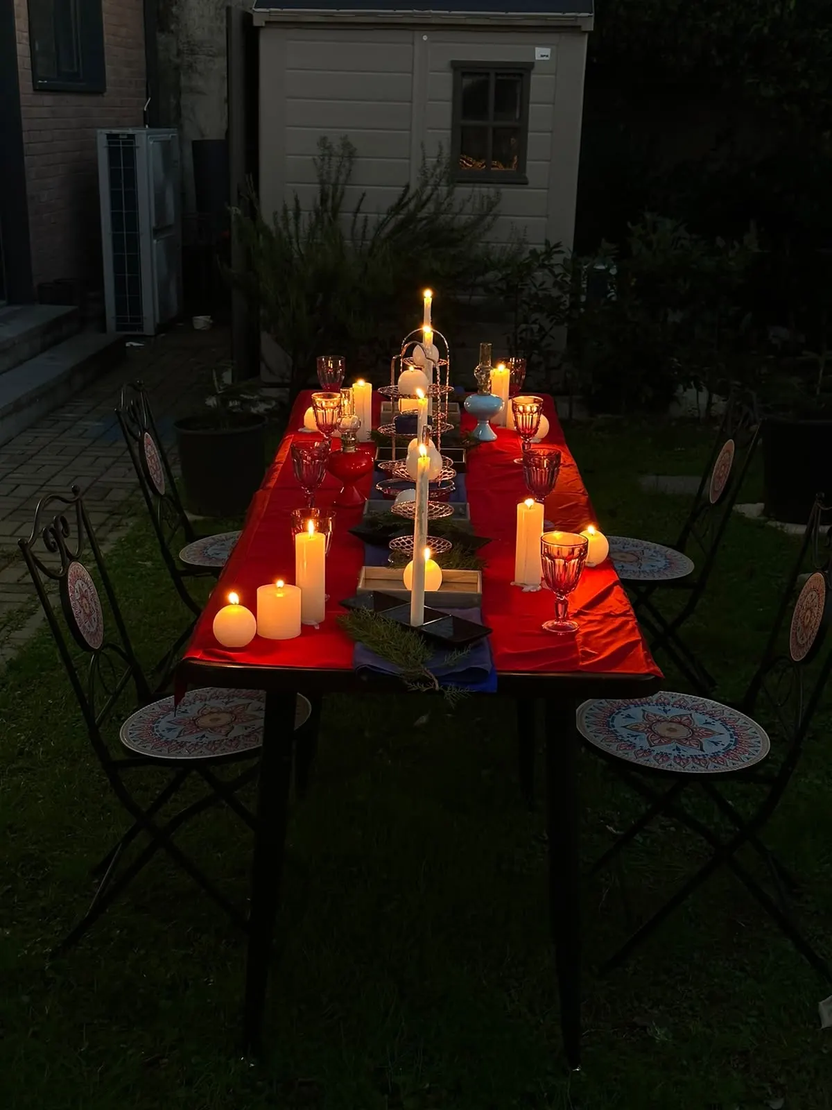 Tall candles glowing on a red table set for a garden dinner